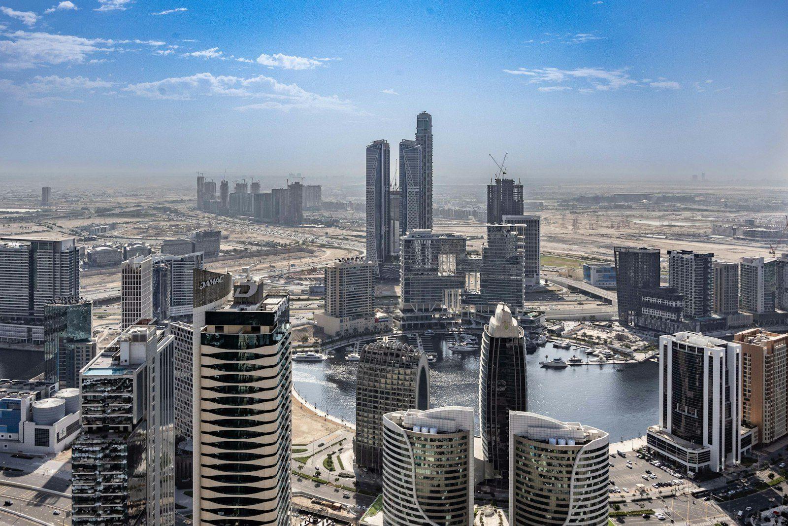 Aerial view of the Business Bay high-rise cluster along the Dubai Water Canal, boats moored at the waterfront and construction cranes among the towers.