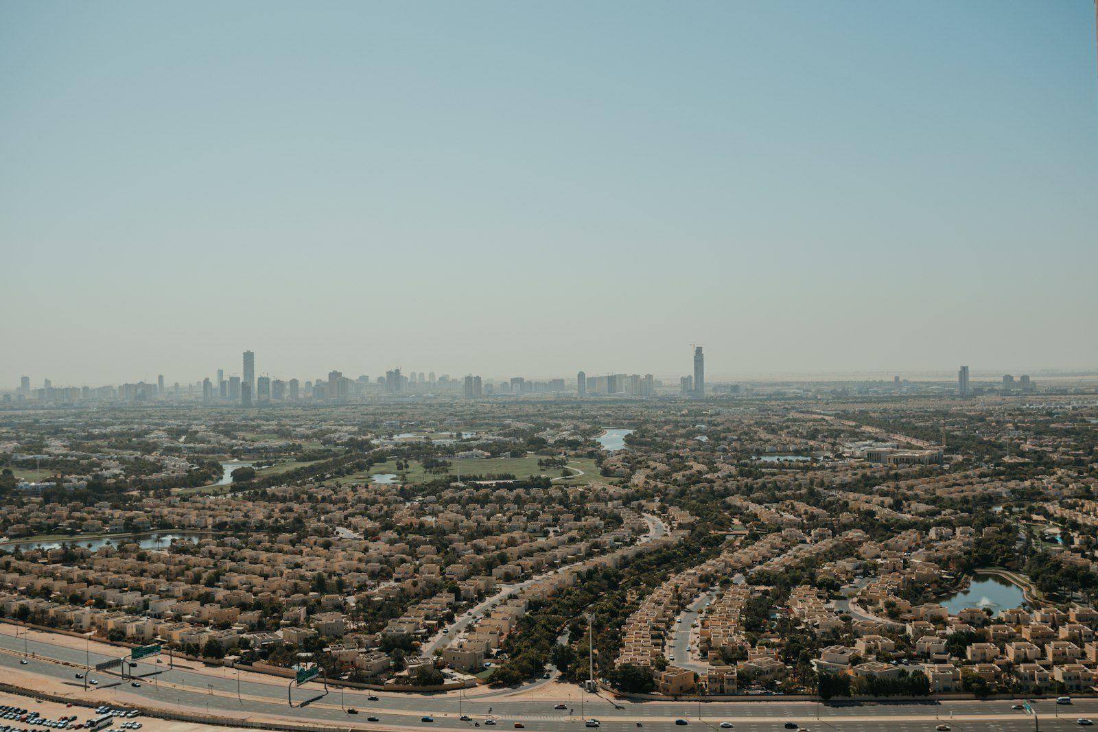Aerial view across a low-rise Dubai villa and townhouse community, the high-rise city skyline visible on the hazy horizon.