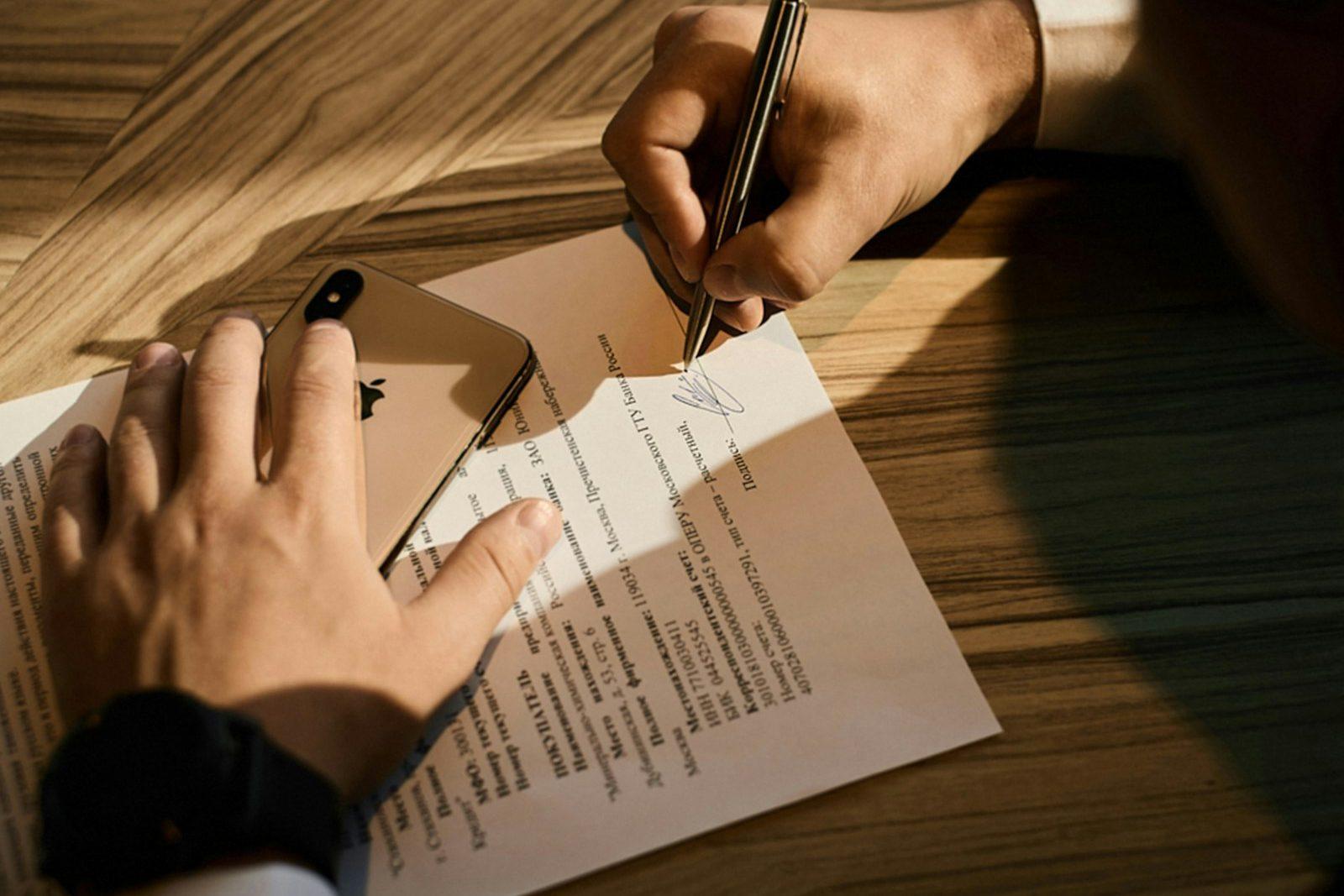 Two people at a desk in raking light, one signing a printed agreement while the other holds the page steady.