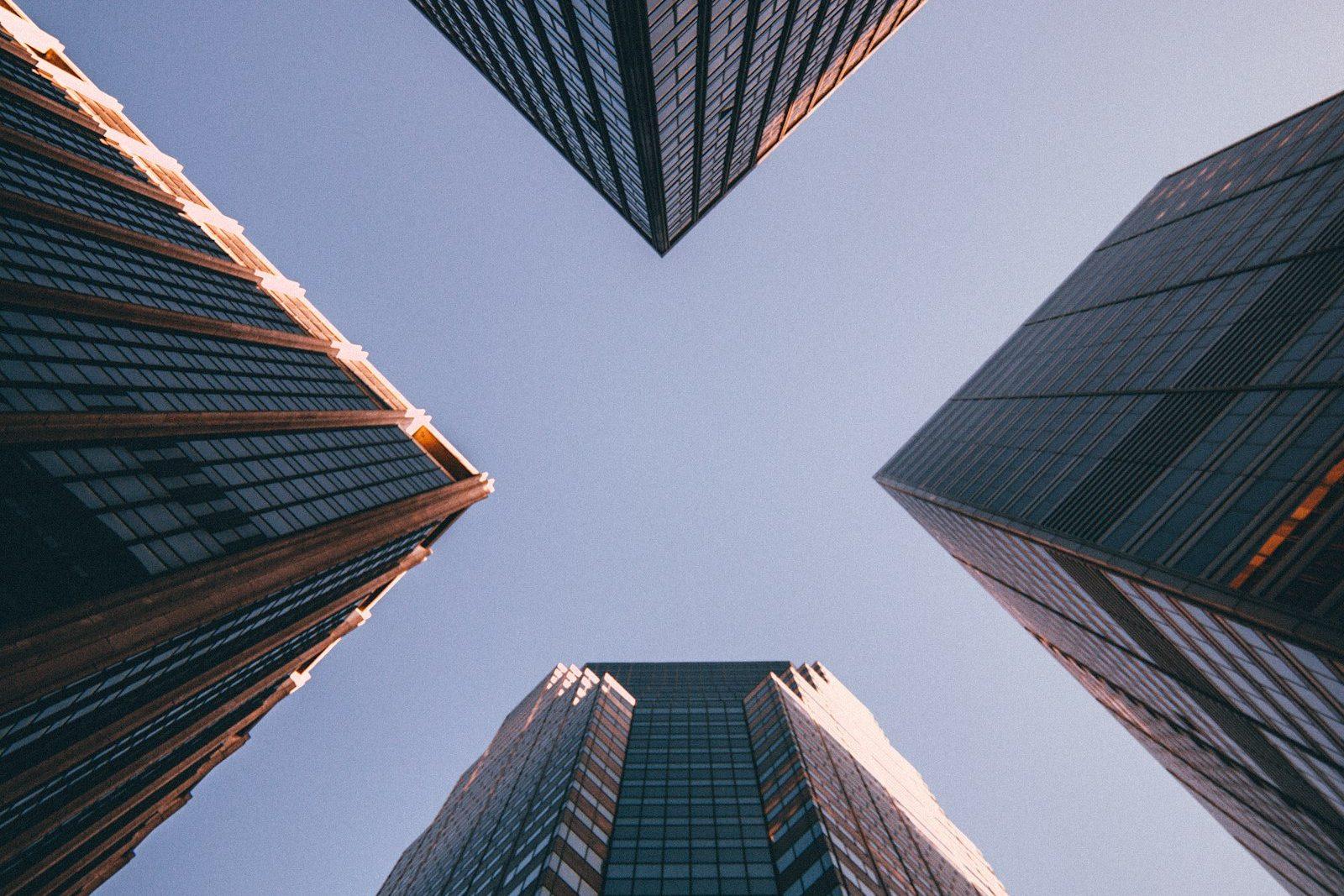Looking straight up between four glass office towers, their facades converging on a pale sky.