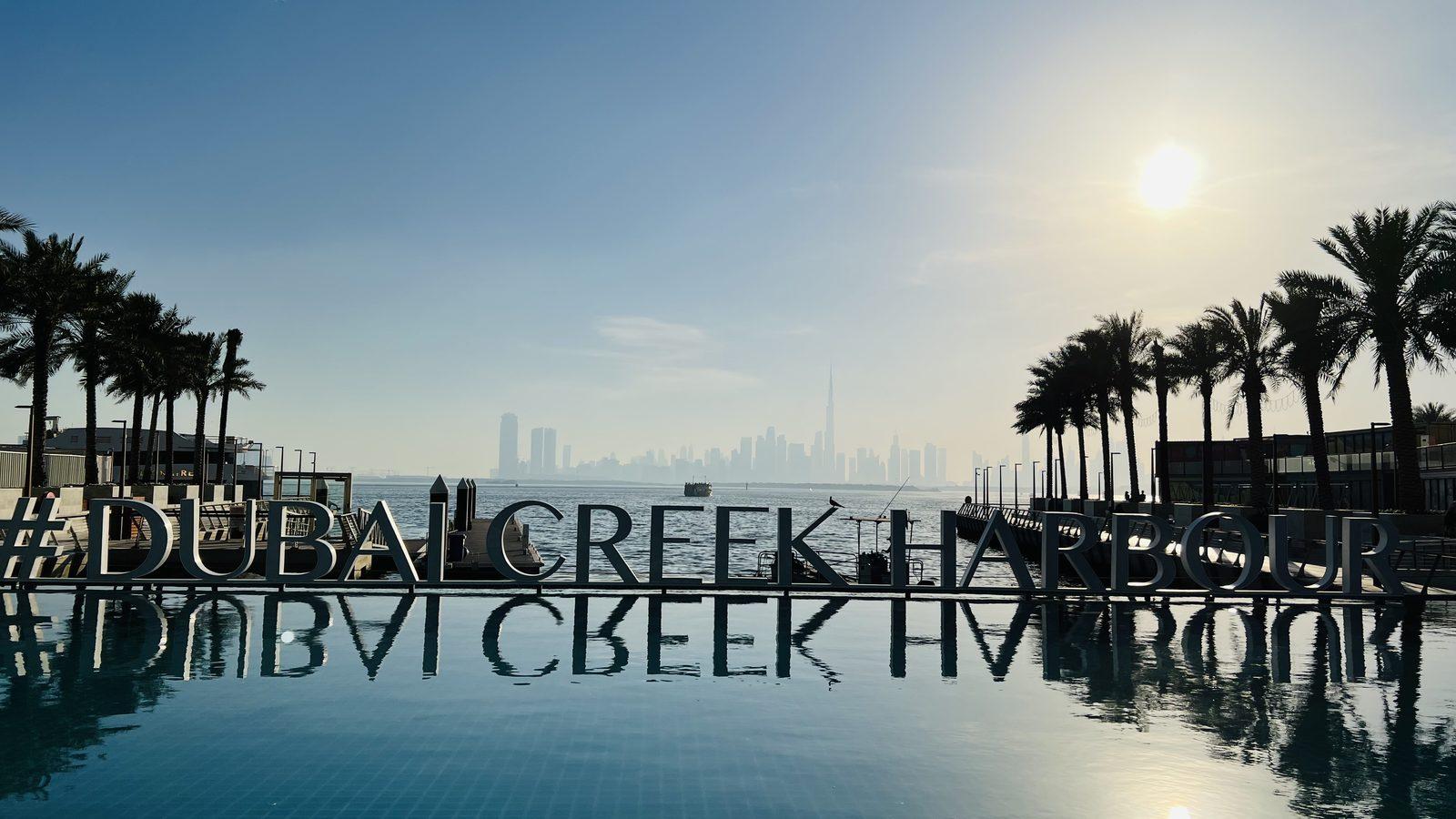 The #DUBAICREEKHARBOUR letter sign standing in a still reflecting pool on the waterfront promenade, date palms to either side, the creek beyond and the Downtown skyline with the Burj Khalifa on the far horizon.