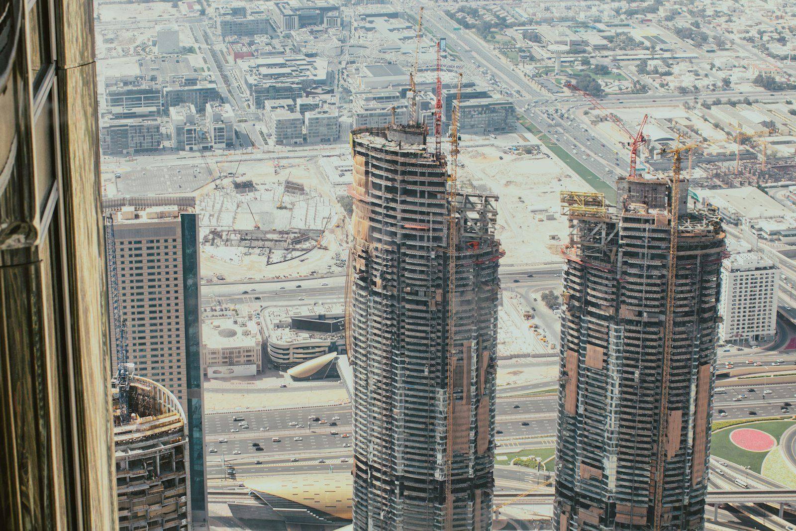 Two residential towers under construction seen from a neighbouring high floor: bare concrete floorplates, tower cranes at the crown, cleared sand plots and further cranes across the district, with traffic on the highway below.