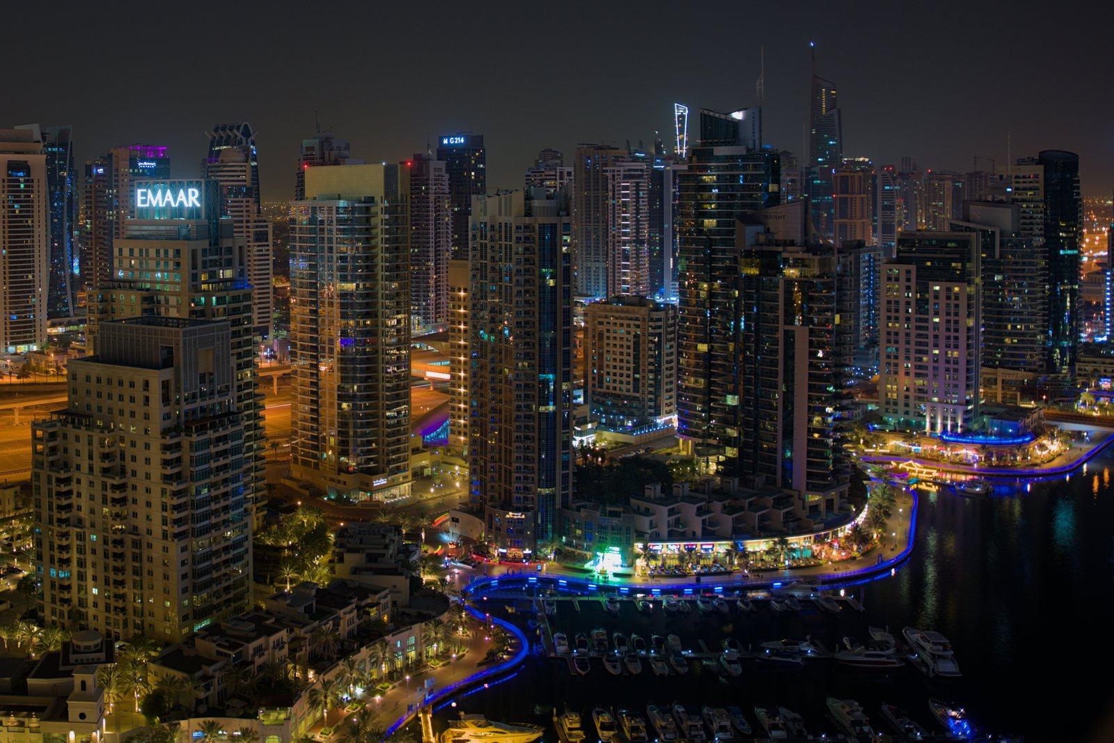 Dubai Marina photographed at night from a high floor: the lit tower cluster packed around the marina basin, motor yachts moored along the quay and the blue-lit curve of Marina Walk tracing the water.