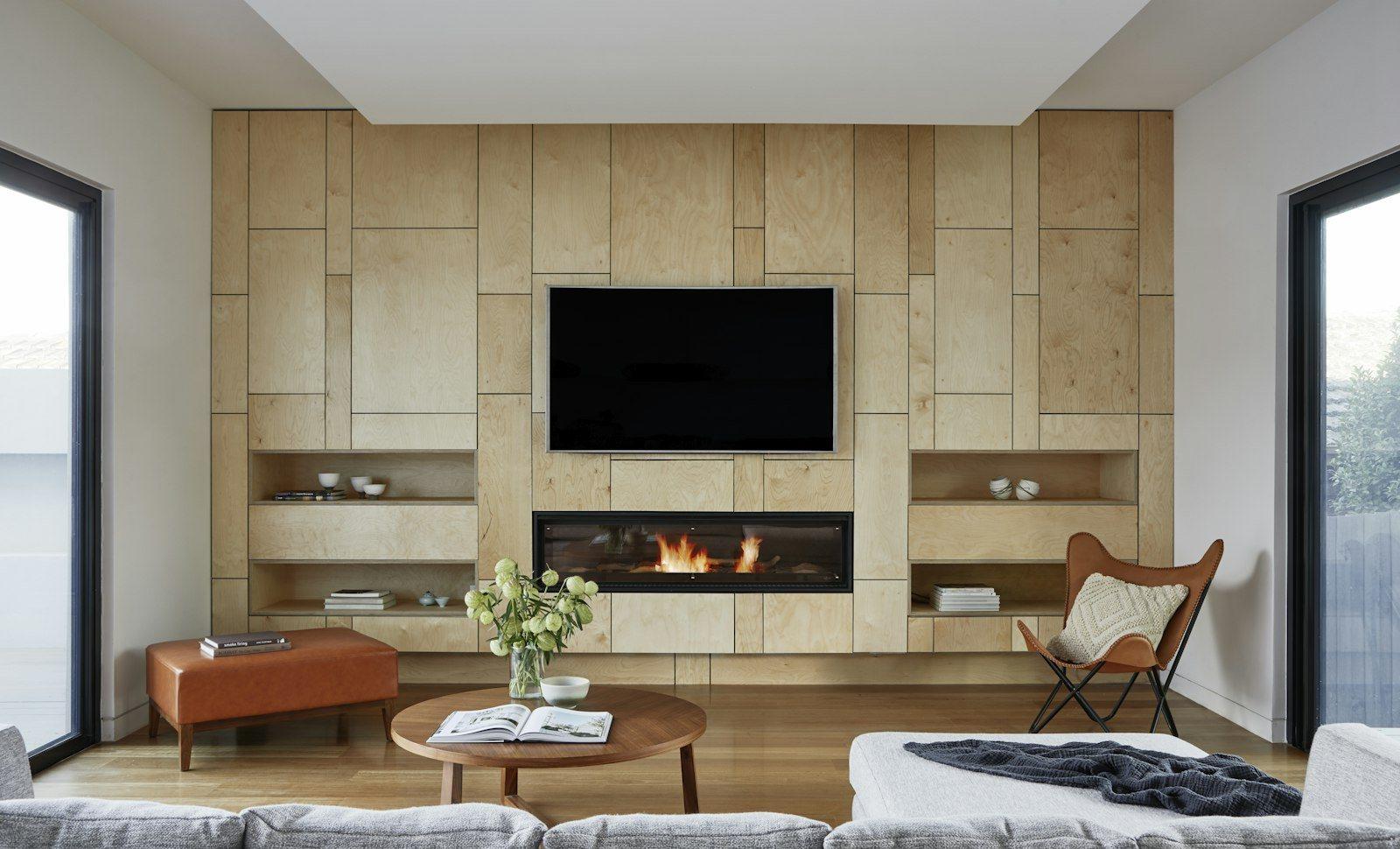 Reception room finished with a travertine feature wall, a long linear gas fire set beneath a wall-mounted screen, a tan leather bench and a low grey sectional, daylight entering through full-height glazing on both sides.