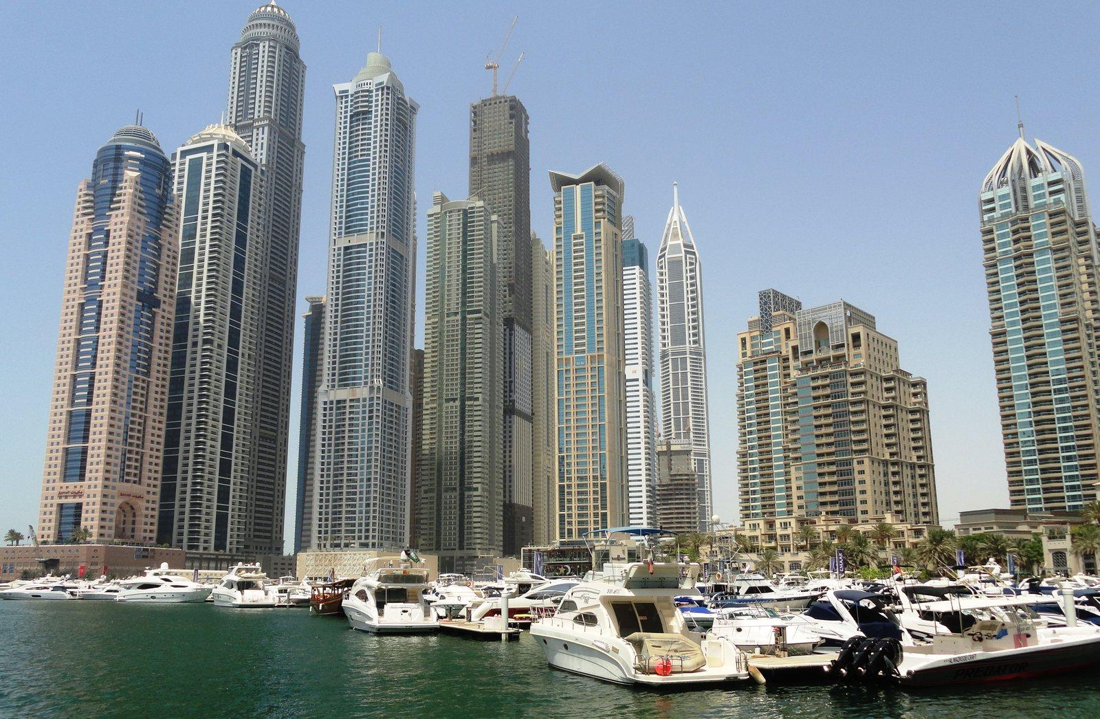 The Dubai Marina tower cluster seen across the water in full daylight, motor yachts berthed along the marina quay in front of the high-rise wall.