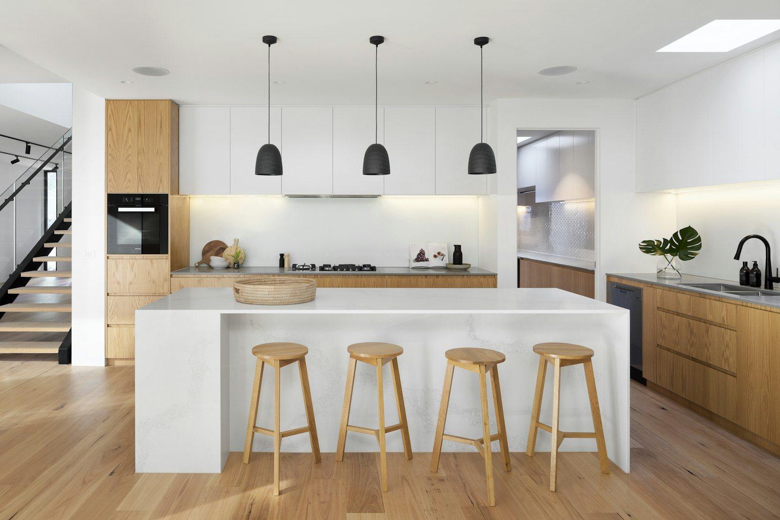 Kitchen with a full-height stone island and four oak stools, pale timber cabinetry, integrated oven and three black pendant lamps, a steel staircase rising at the left.