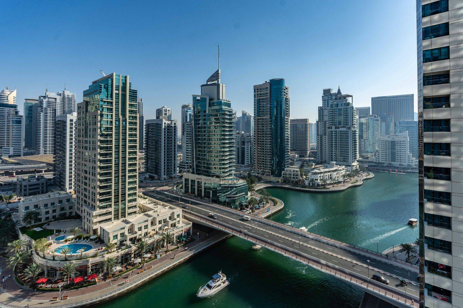 Dubai Marina from a high floor: the waterway curving under the marina bridge, a motor yacht crossing the basin and the Marina Walk promenade lined with cafes below the towers.