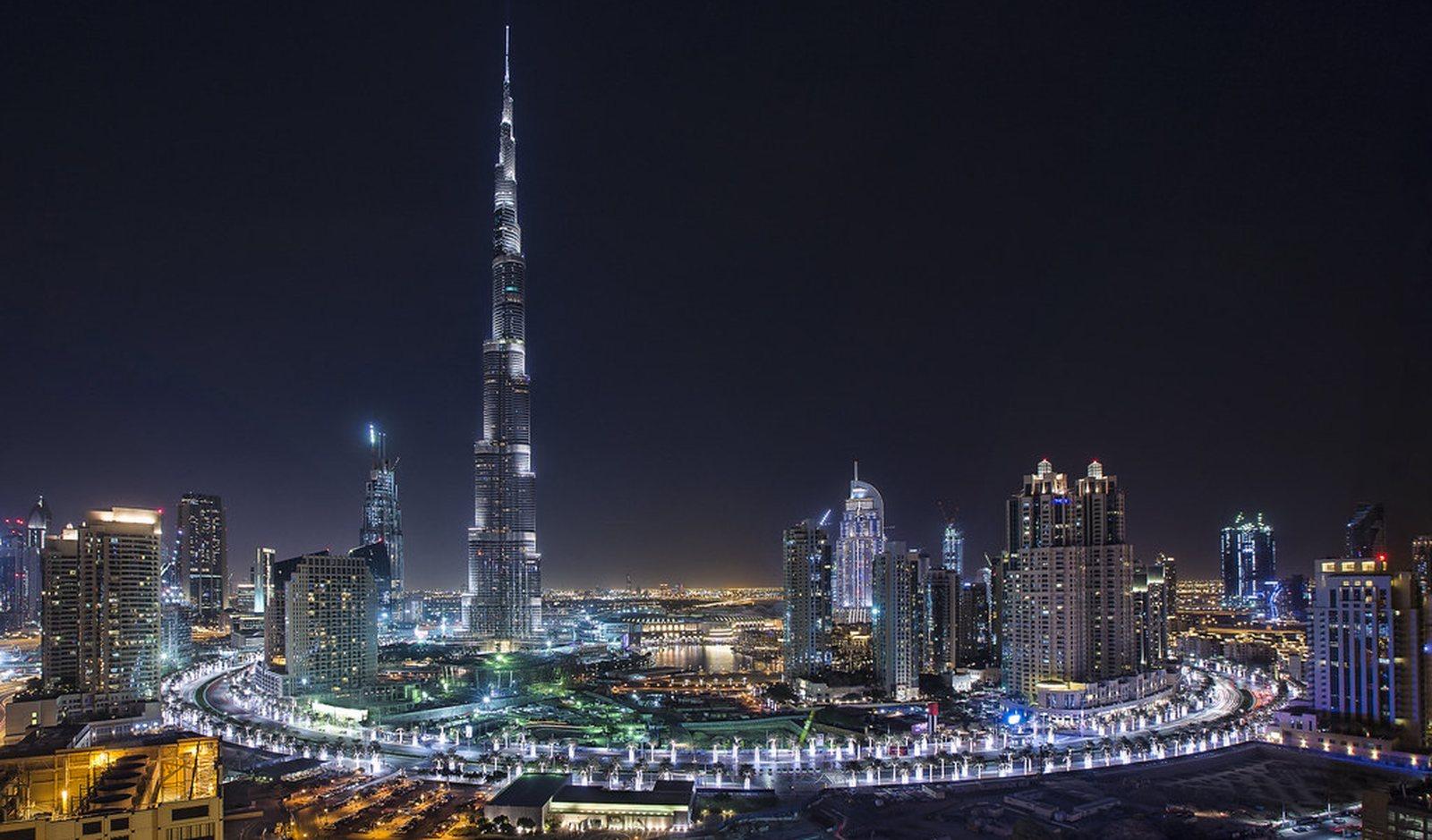 Downtown Dubai at night from a high floor: the Burj Khalifa lit against a black sky, the Dubai Fountain lake at its foot and the ring of Boulevard towers glowing below.