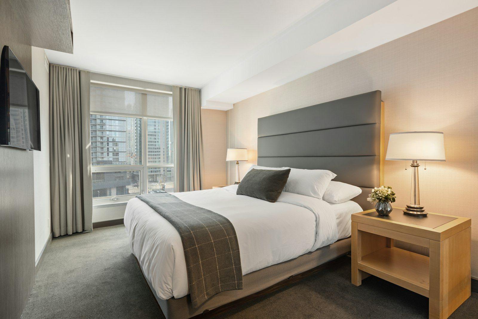 Bedroom with a wide grey upholstered headboard, brass table lamps and a picture window looking straight across at neighbouring residential towers.
