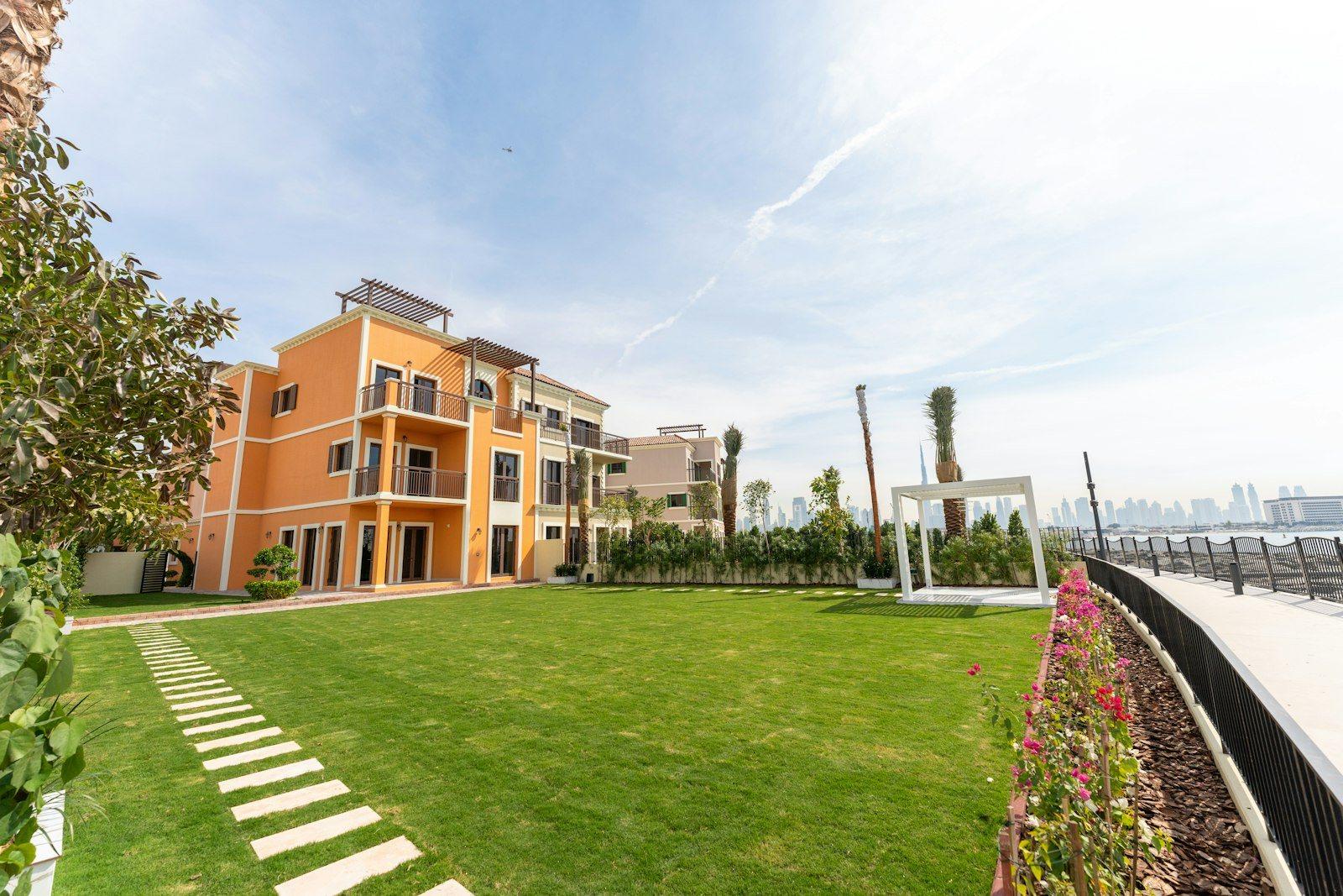 A terrace of townhouses facing a wide lawn with a shaded pergola, a waterfront railing to one side and the Dubai skyline on the horizon.