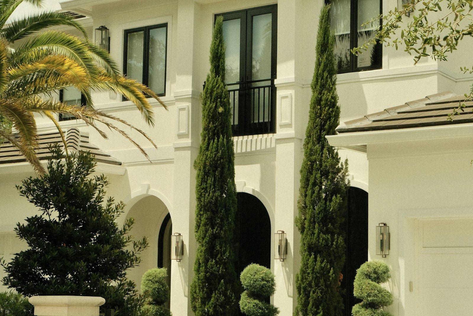 The entrance elevation of a large white villa: an arched, double-height porch flanked by lanterns, clipped cypresses and topiary in stone urns, with a date palm overhanging the drive.