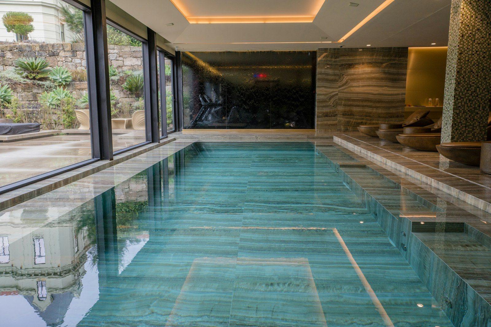 Indoor pool lined in onyx and travertine, a dark water wall at the far end and sliding glazing onto a planted terrace.