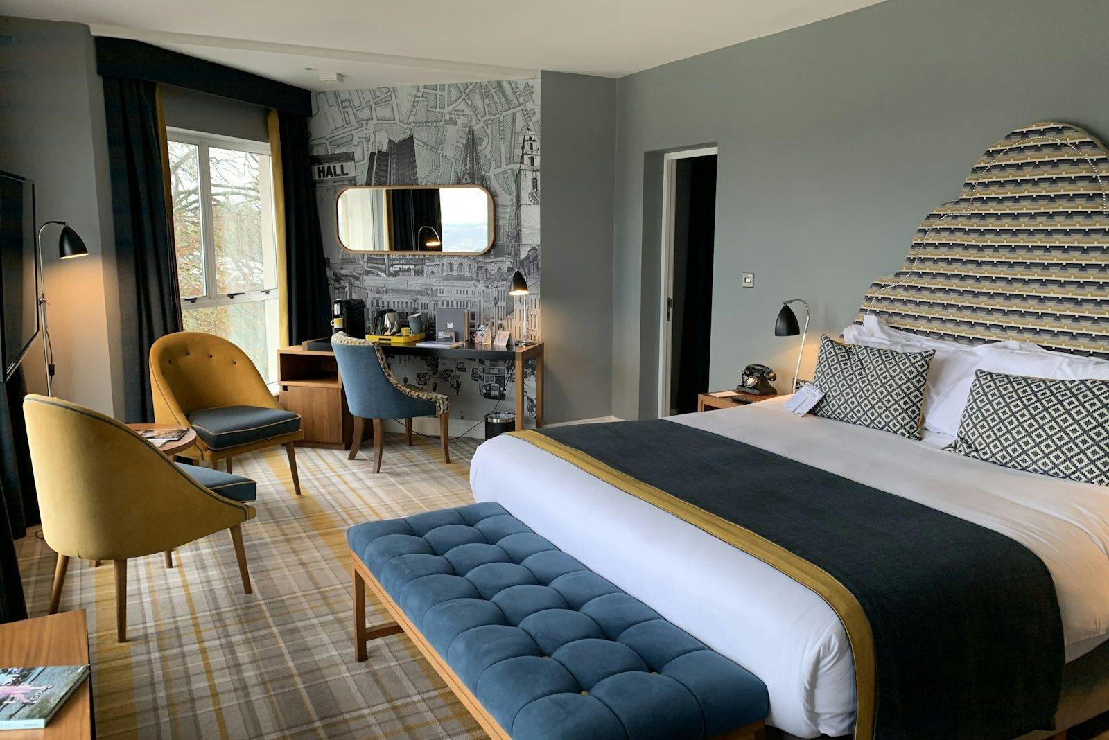 Bedroom with a fan-pleated patterned headboard, a blue upholstered bench at the foot of the bed, mustard armchairs by the window and a mirrored dressing corner.