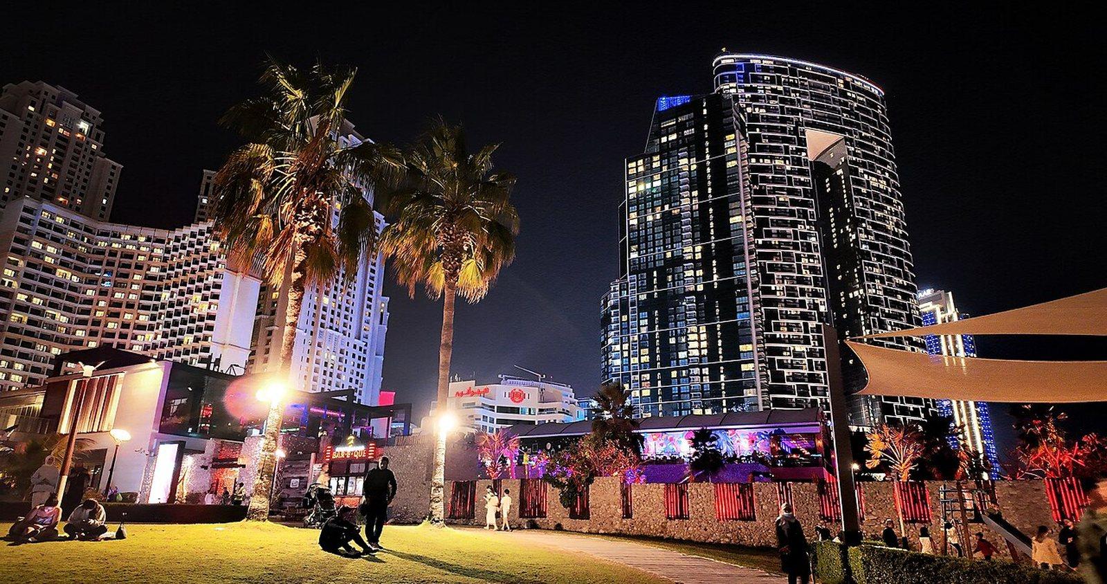 The Walk at Jumeirah Beach Residence after dark, floodlit palms and illuminated retail terraces below a dark curved residential tower.