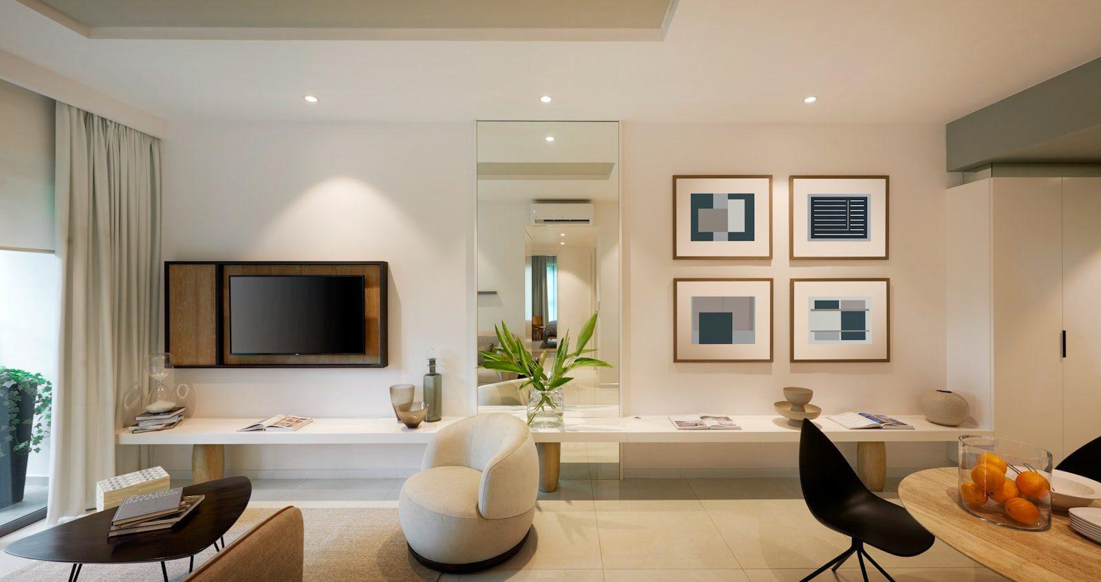 Open-plan living and dining room in a modern apartment: white walls and large pale porcelain floor tiles, a wall-mounted television set into a timber surround, a full-height mirror reflecting a split air-conditioning unit in the hall, a grid of four framed abstract prints, a cream bouclé tub chair and a round timber dining table.