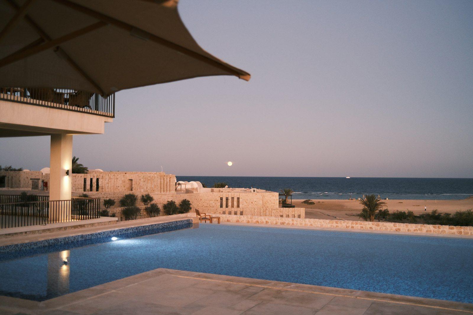 Infinity pool terrace at dusk under a shade canopy, the water reaching towards a sand beach and the Gulf with a low stone building alongside.
