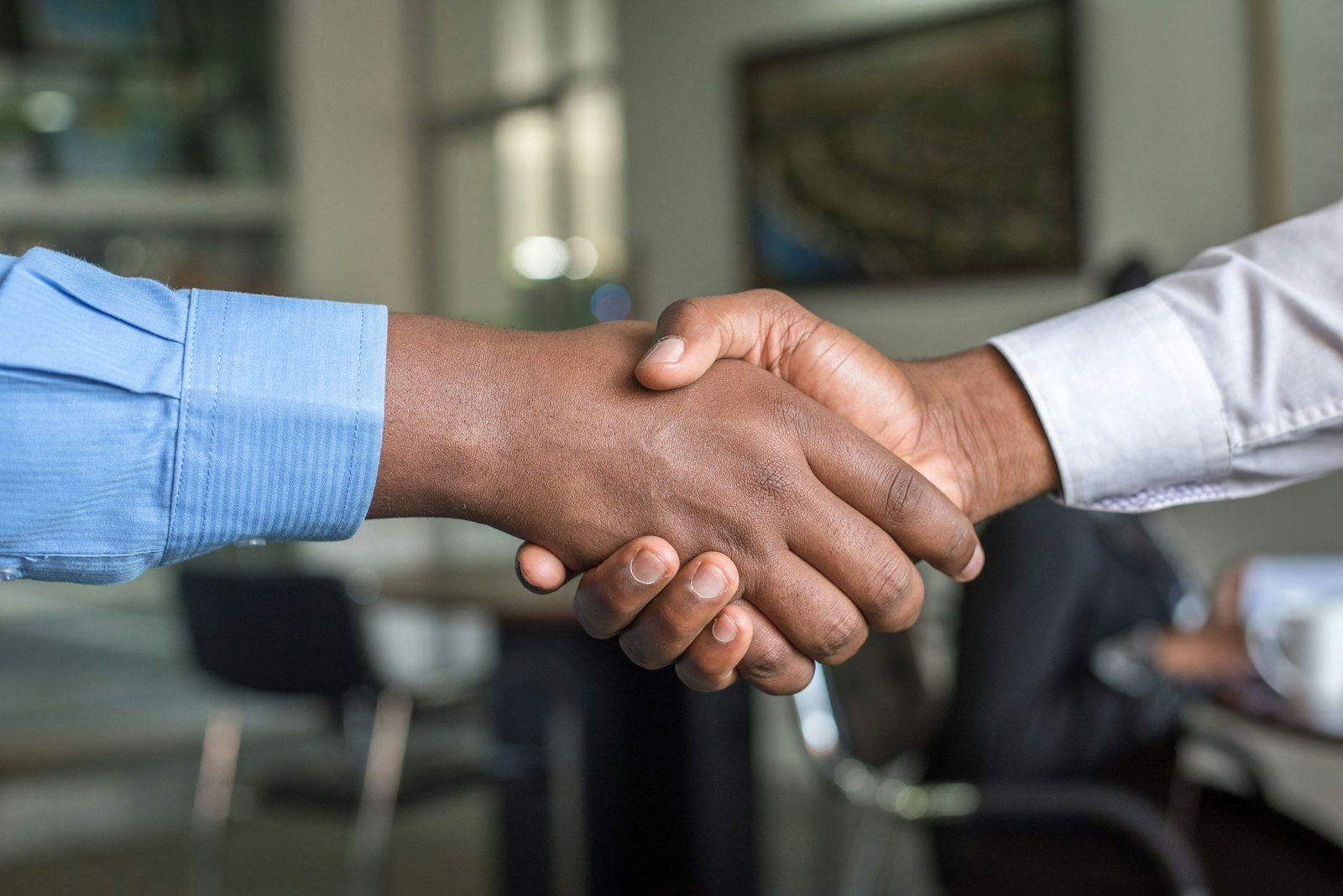 Two people shaking hands in close-up, a meeting room softly out of focus behind them.