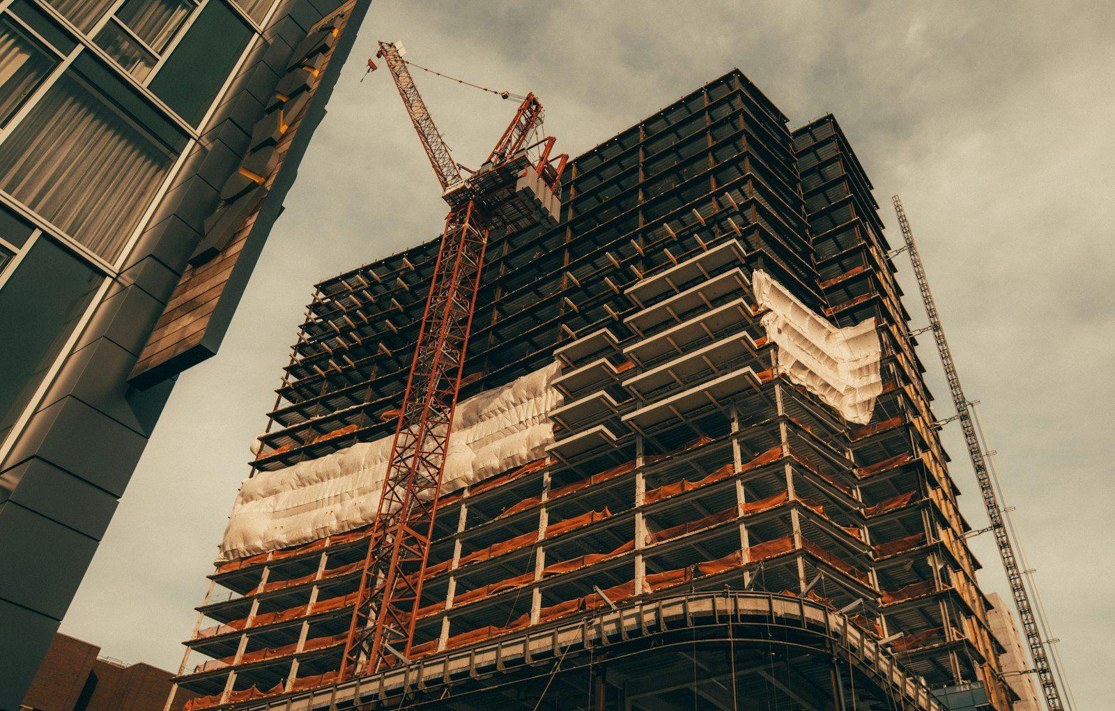 A tower under construction seen from street level, its exposed floor slabs wrapped in netting with a red tower crane rising alongside.
