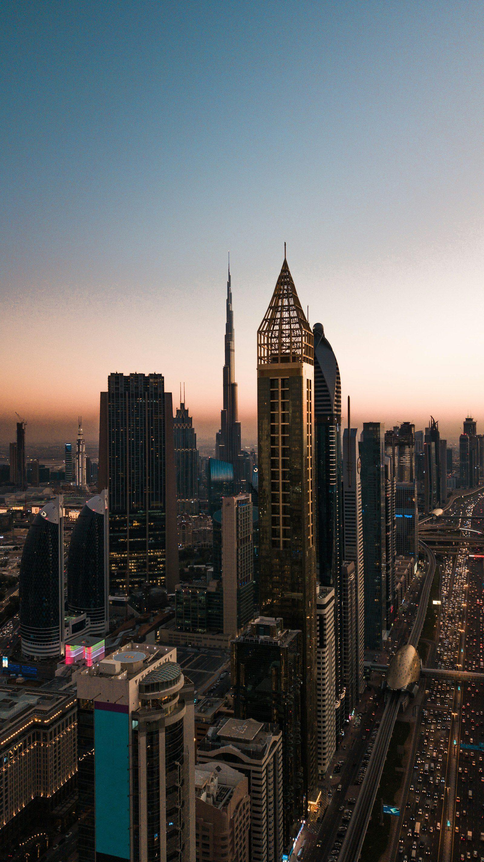 Aerial view along Sheikh Zayed Road at dusk, the Burj Khalifa lit against a deep orange horizon with traffic threading between the towers below.