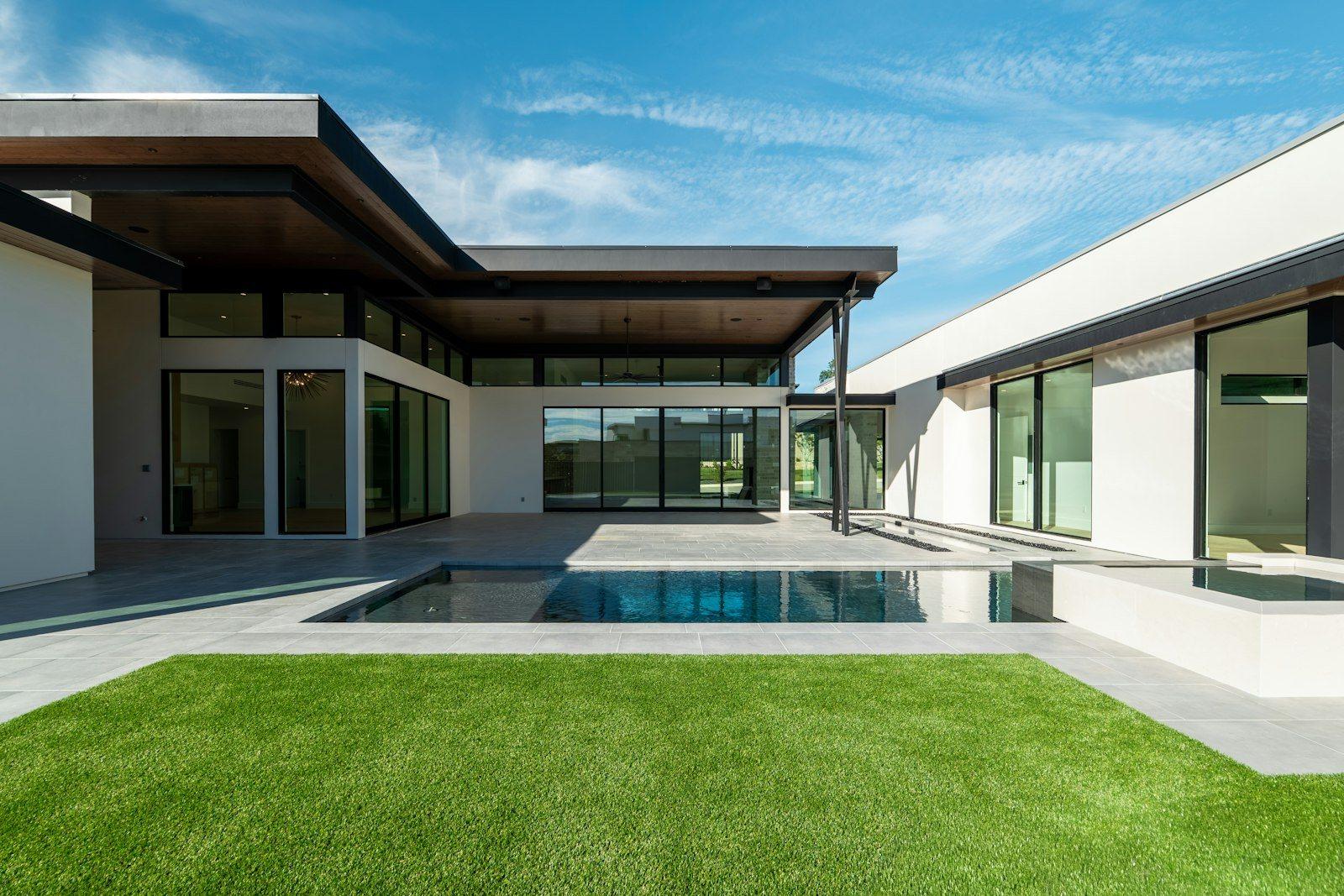 The same villa from the pool terrace, the glazed living pavilion set back behind a shaded loggia with the lawn wrapping around it.