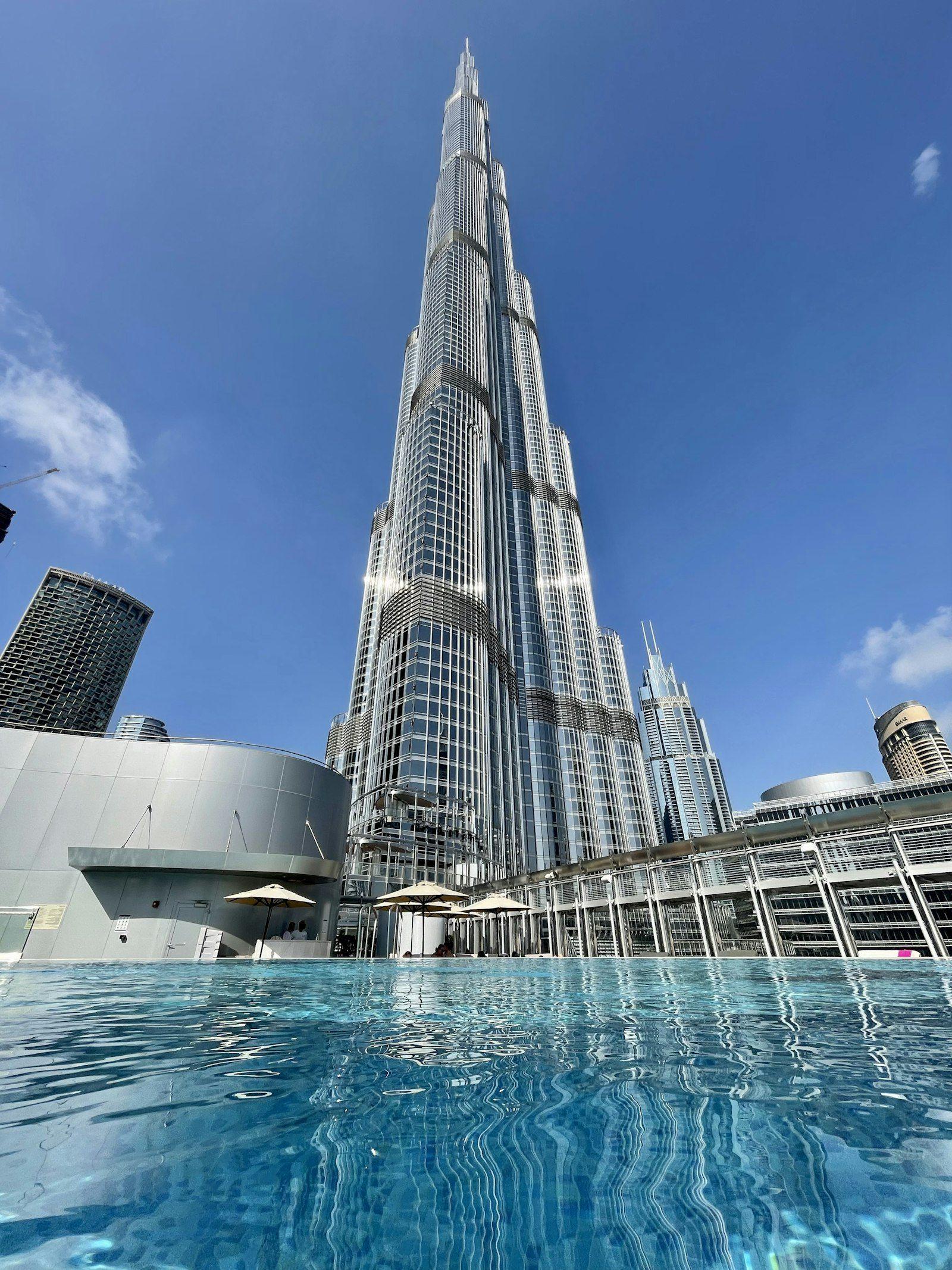 The Burj Khalifa rising above a resident pool deck, seen from the waterline with parasols and loungers along the far side and the Downtown towers to the right.
