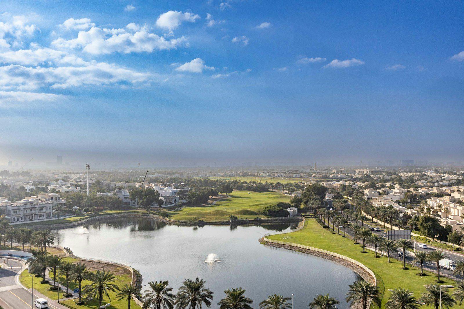 A golf community from the air: a fairway and green running between palm-lined roads, an irrigation lake with fountains in the foreground and large villas backing onto the course on both sides.