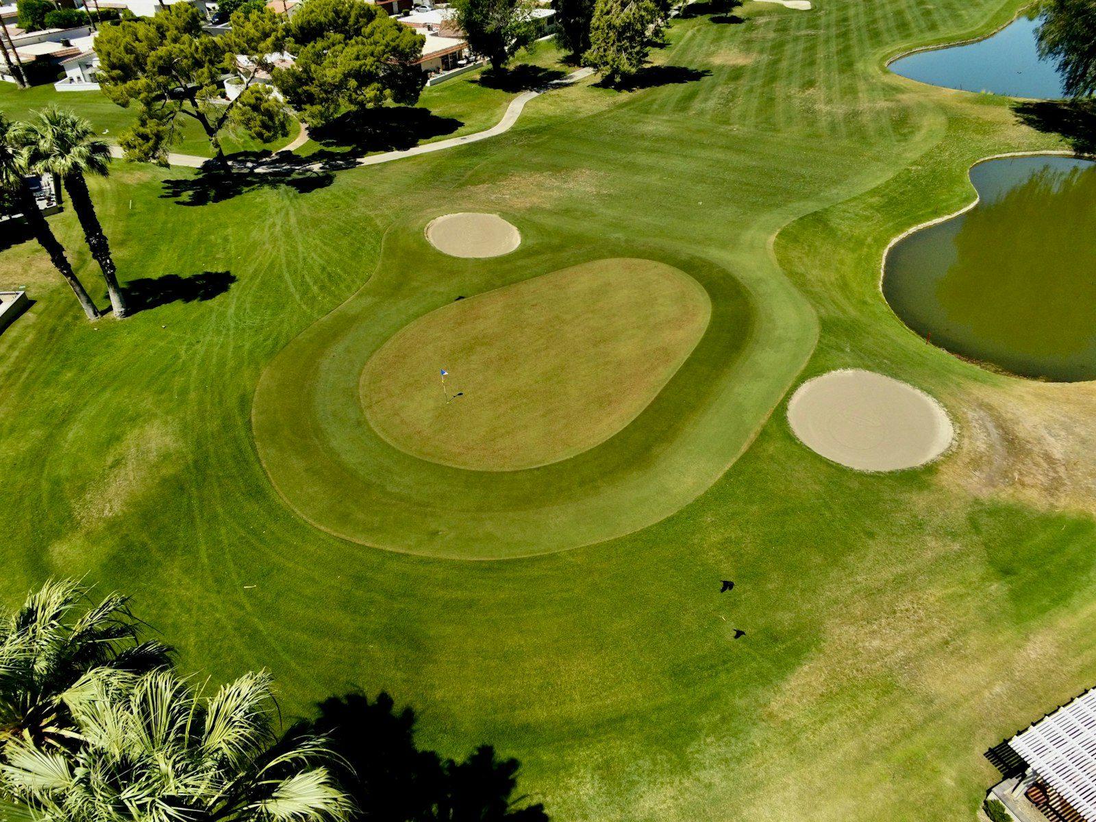 Aerial view of a golf green ringed by bunkers and a water hazard, palms along the cart path and houses backing directly onto the fairway.