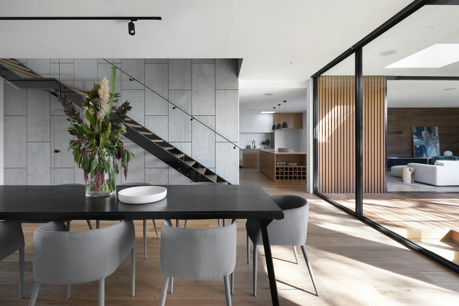 Double-height dining area of a duplex: a black oval table with grey chairs, a steel stair rising against a concrete wall and an open kitchen and sitting room beyond the glazed partition.