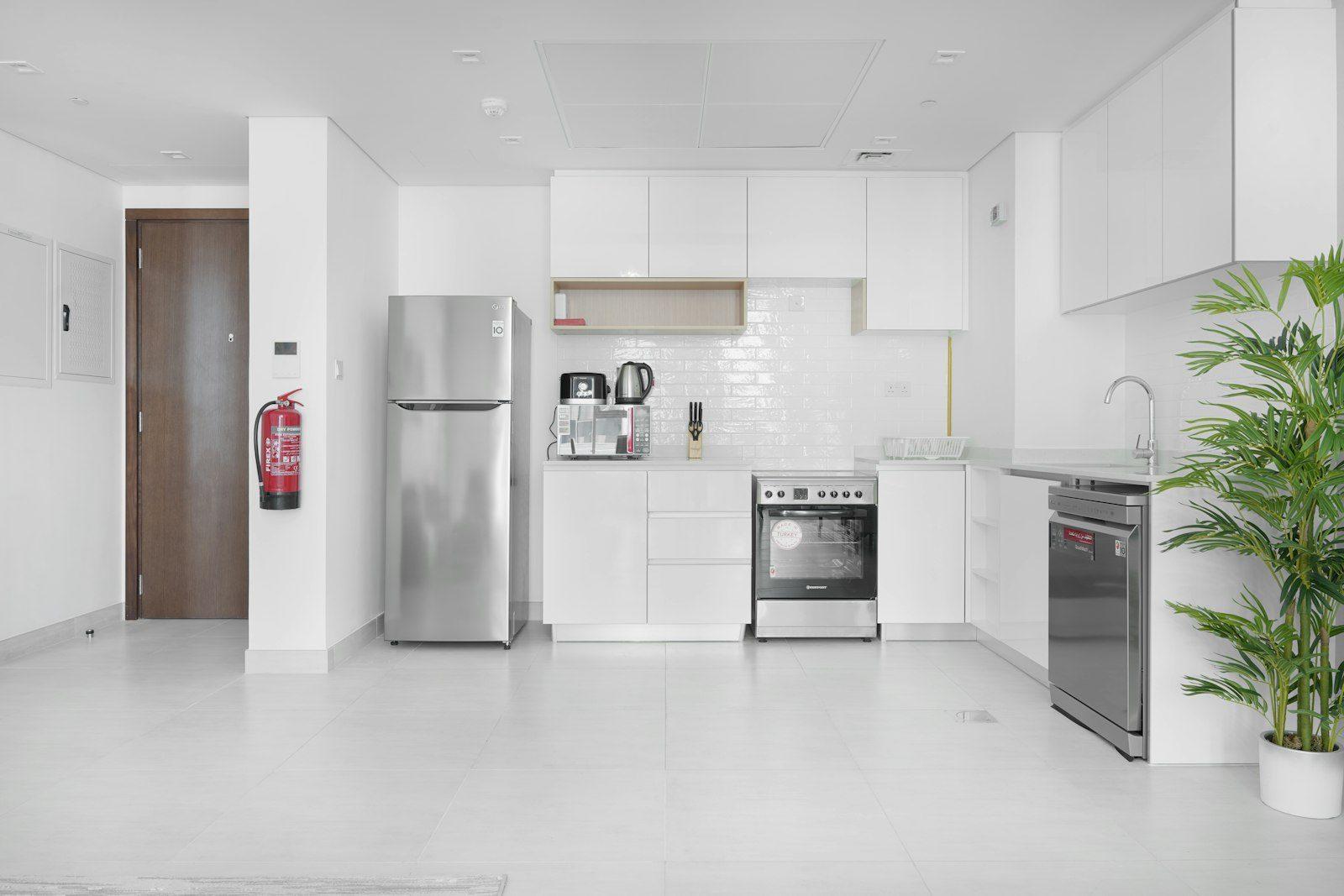 Apartment kitchen finished in white handleless gloss units with a white metro-tile splashback, a freestanding cooker, dishwasher and fridge-freezer, a ceiling air-conditioning return grille and a red dry-powder extinguisher beside the timber entrance door.