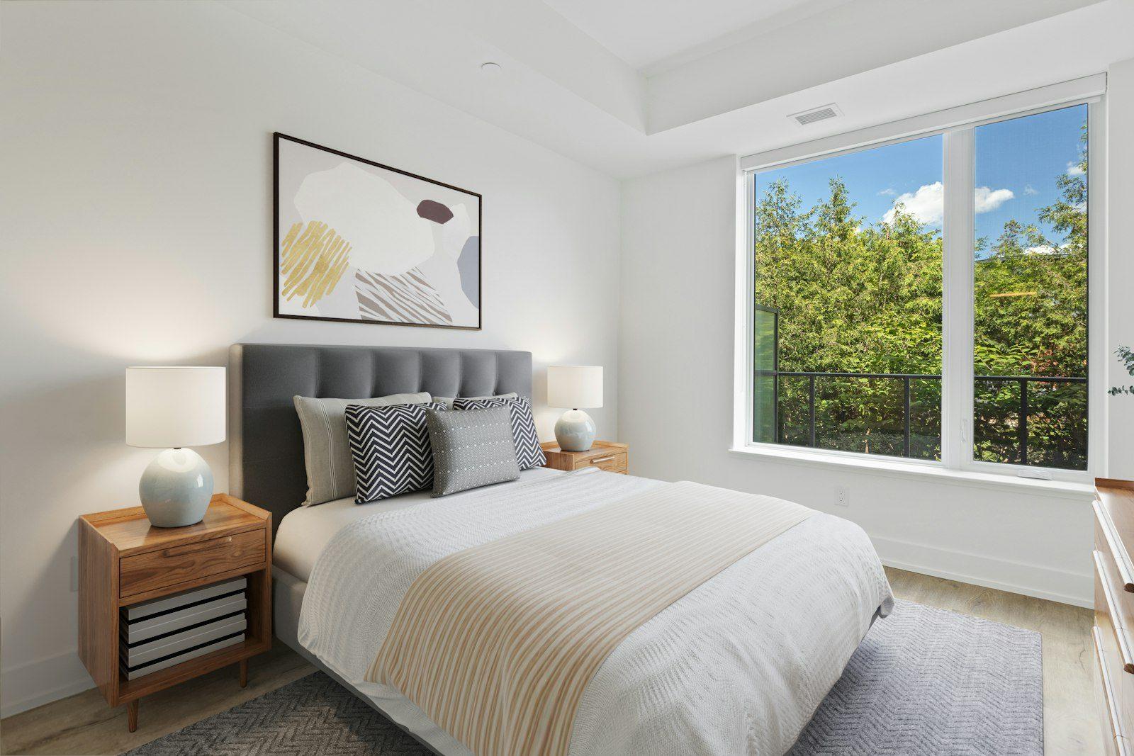 Bedroom with a grey buttoned headboard, timber bedside tables and ceramic lamps, an abstract canvas above the bed and a wide window with a Juliet balcony looking into mature green trees.
