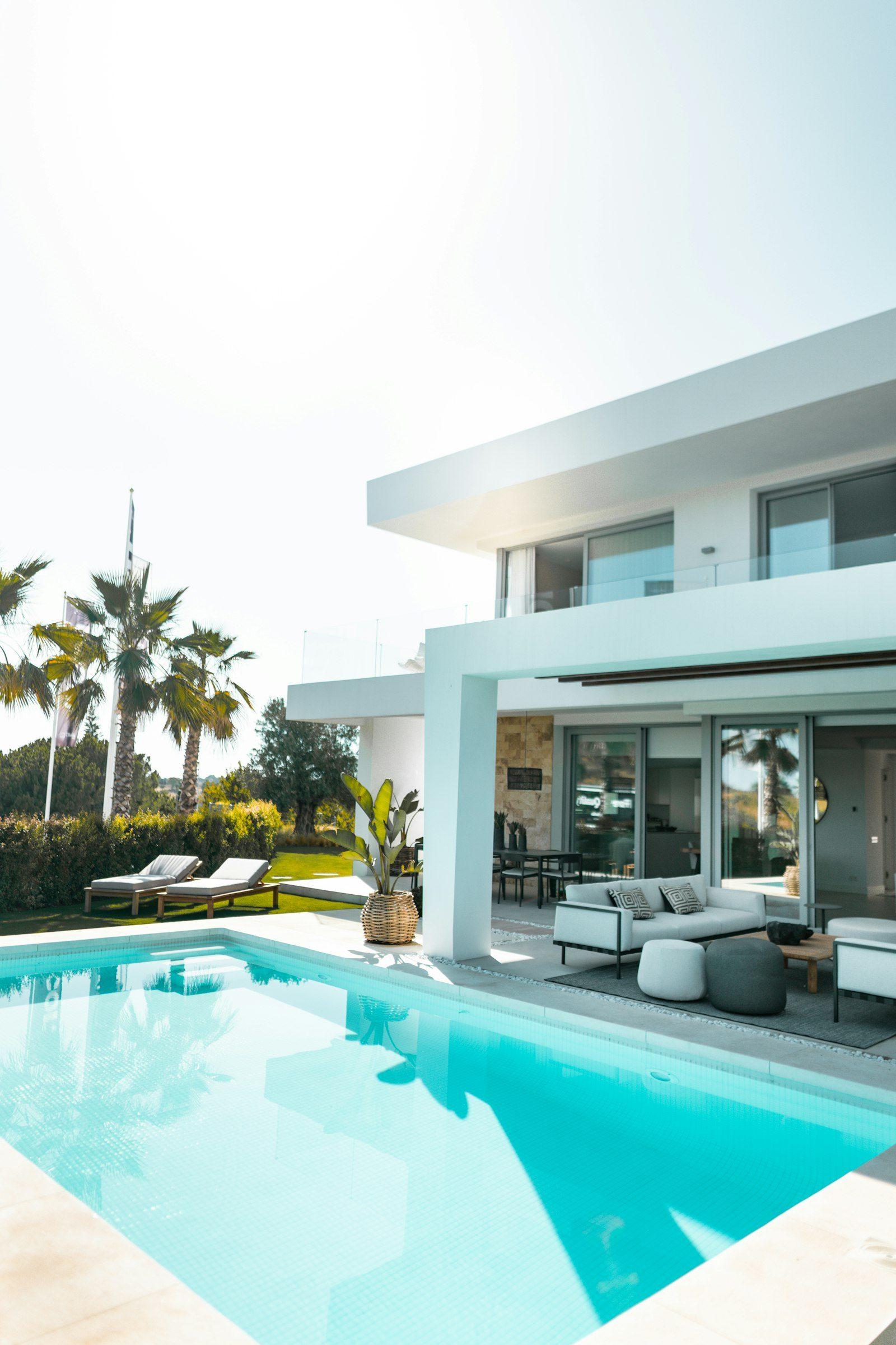 Villa pool terrace: a turquoise mosaic-tiled pool beside a shaded loggia furnished with a pale outdoor sofa and dining table, two loungers on the lawn and palms along the boundary.