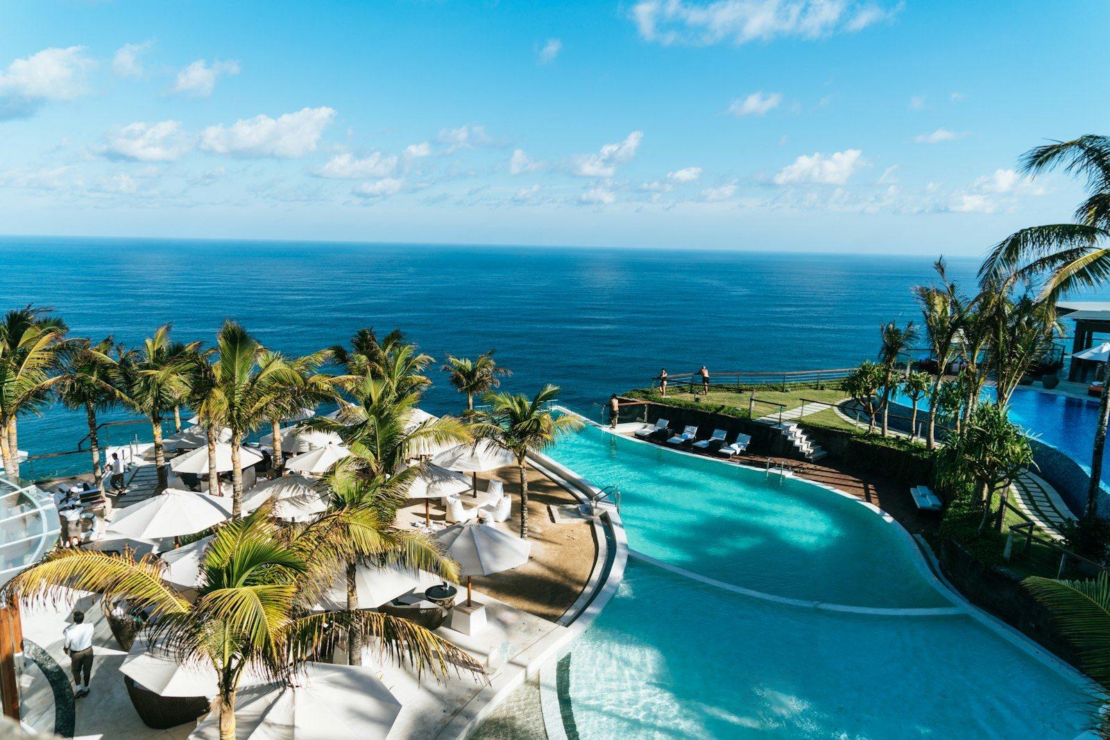 A five-star beach club above a deep blue sea: a wide free-form pool edged with palms, white parasols and loungers, and a second infinity pool on the terrace beyond.