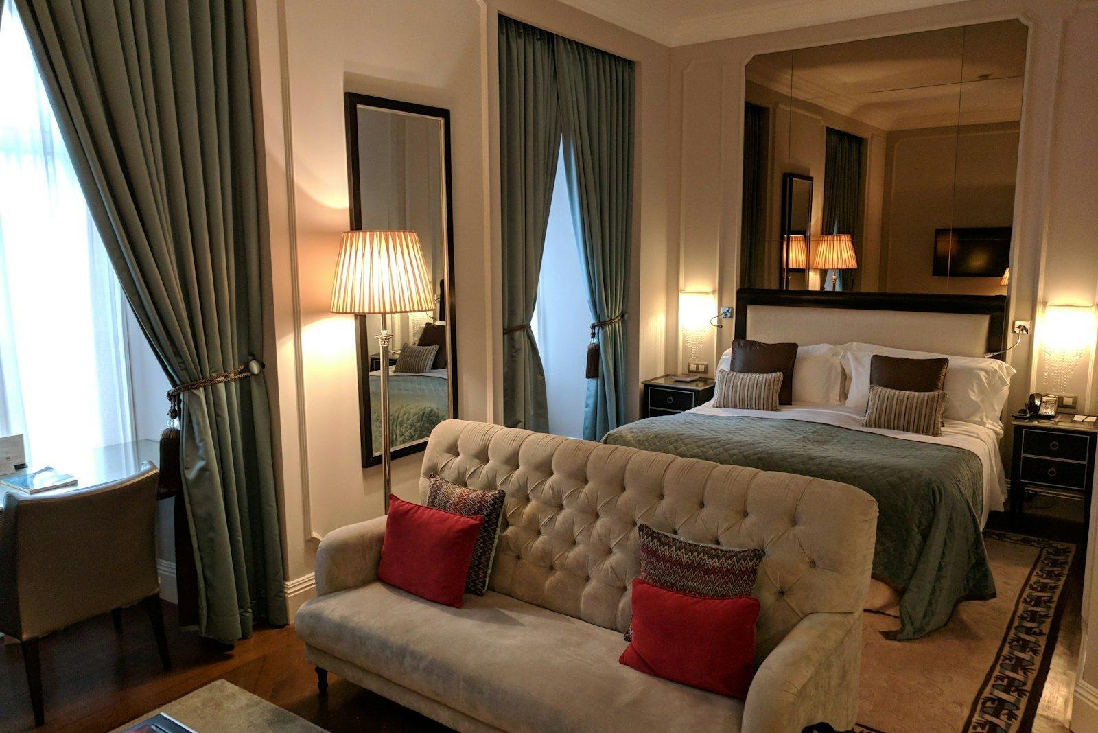 Suite bedroom in a classical register: olive silk drapes, mirrored wall panels, a buttoned sofa at the foot of the bed and lamplit bedside recesses.
