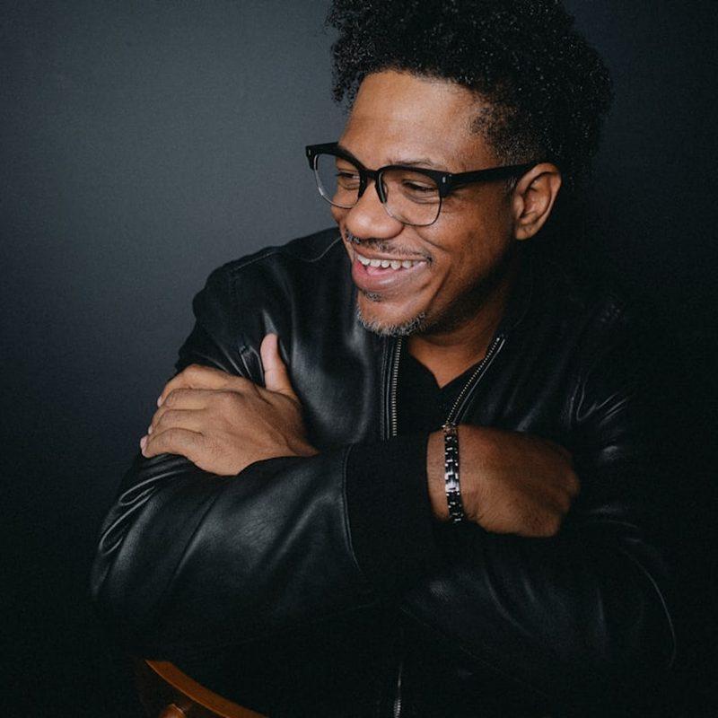 Portrait of a man in glasses and a dark jacket, arms folded, against a dark studio backdrop.