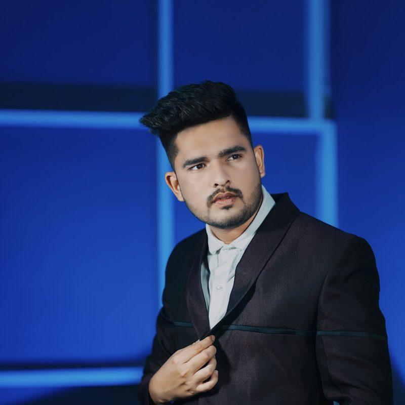 Portrait of a man in a black suit adjusting his jacket, lit against a deep blue studio backdrop.